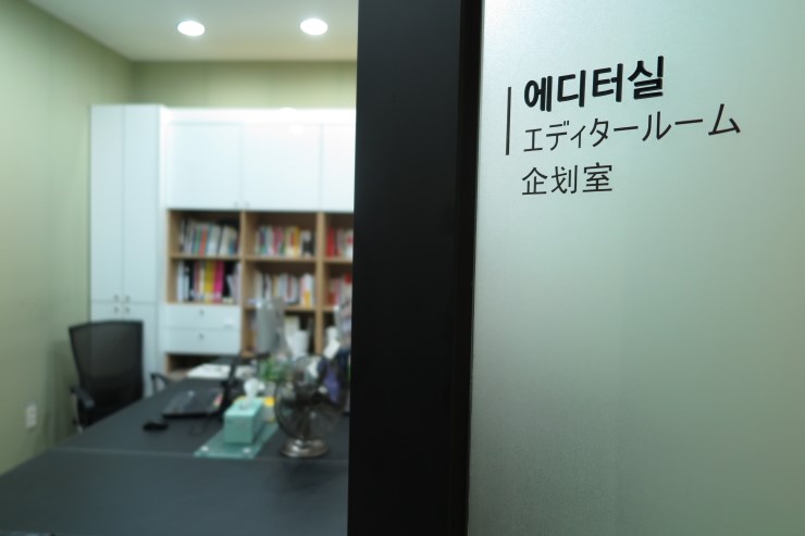 에디터실 문이 살짝 열려있고, 책이 꽂힌 책장이 보이고 업무를 하는 책상이 보인다.