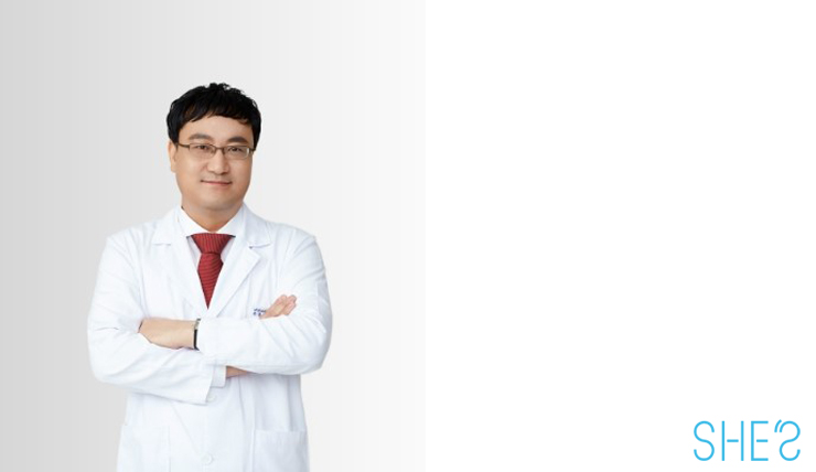의사가운을 입고 팔짱을 끼고 정면을 주시하며 옅은 미소를 짙고 있다